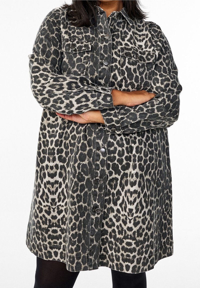 Personne portant une veste à manches longues imprimé léopard boutonnée, les bras croisés, debout devant un fond blanc uni.