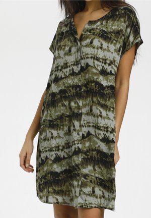 Woman wearing a short-sleeve, button-up shirt dress with green and black tie-dye pattern, standing with one hand in pocket against white background.