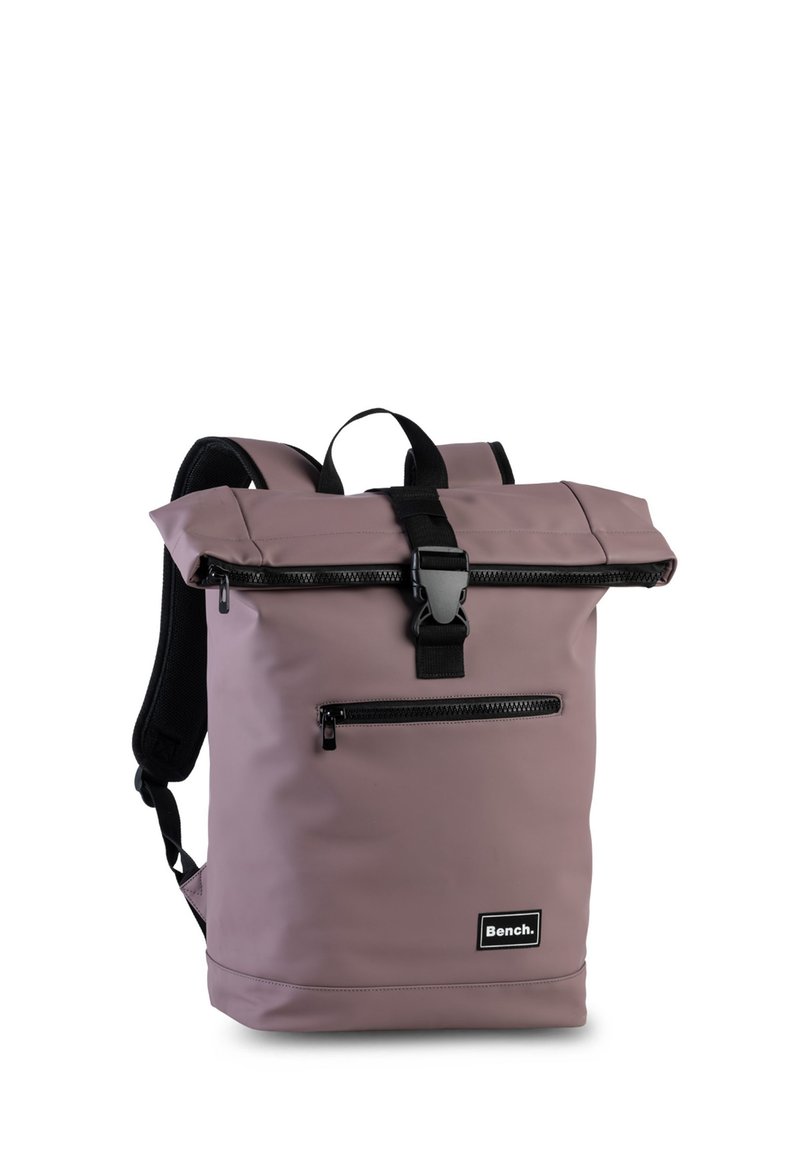 Mochila morada tipo roll-top hecha de material duradero con correas negras. Presenta un bolsillo frontal con cremallera y un parche con el logo en la parte inferior.