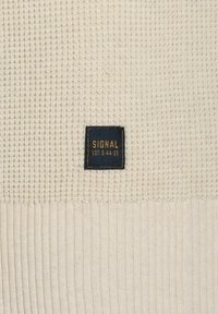 Strickpullover in Cremefarbe, mit strukturiertem Oberteil und geripptem Saum. Schwarzes Etikett mit der Aufschrift "SIGNAL" in goldener Schrift für das Branding.
