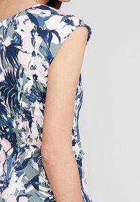 Femme portant une robe sans manches à motif floral bleu, rose et blanc, vue de dos et de profil sur un fond gris clair.