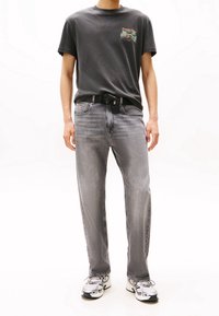 Man wearing black graphic t-shirt, grey faded jeans with black belt, and white sneakers on white background, standing facing forward.