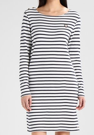 Vrouw draagt een witte jurk met lange mouwen en zwarte horizontale strepen, met een klein hartvormig plaatje op de borst, staand tegen een effen achtergrond.