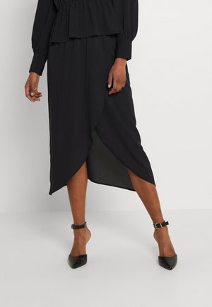 Mujer con top de manga larga negro y falda midi negra asimétrica, con tacones negros de punta cerrada y correa en el tobillo, posando frente a un fondo blanco.