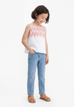 Jeune fille aux cheveux courts et foncés portant un débardeur blanc avec un motif zigzag rose, un jean bleu clair et des sandales beige, les mains dans les poches.