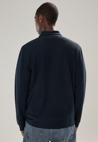 Námornícka modrá polokošeľa s dlhým rukávom, golierom a textúrovanou látkou. Model nosí svetlomodré rifle. Pohľad je zozadu.