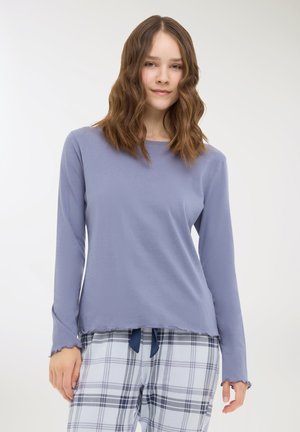 Giovane donna con capelli castani ondulati che indossa una maglietta a maniche lunghe di un delicato colore viola e pantaloni del pigiama bianchi a quadri con cordoncino blu navy.