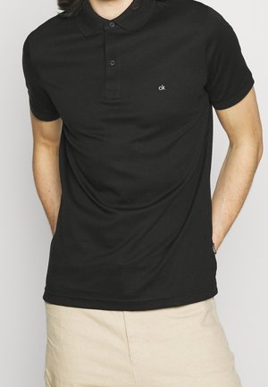 Muž v černém krátkém polo tričku s logem "ck" na hrudi a béžových šortkách, ruce v kapsách, na jednobarevném pozadí.