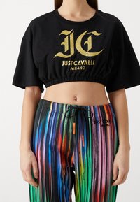 Mustavihreä cropped t-paita, jossa on kultaglitterinen logo "JUST CAVALLI MILANO", yhdistettynä monivärisiin raidallisiin housuihin, joissa on nyörikiinnitysvyötärö.