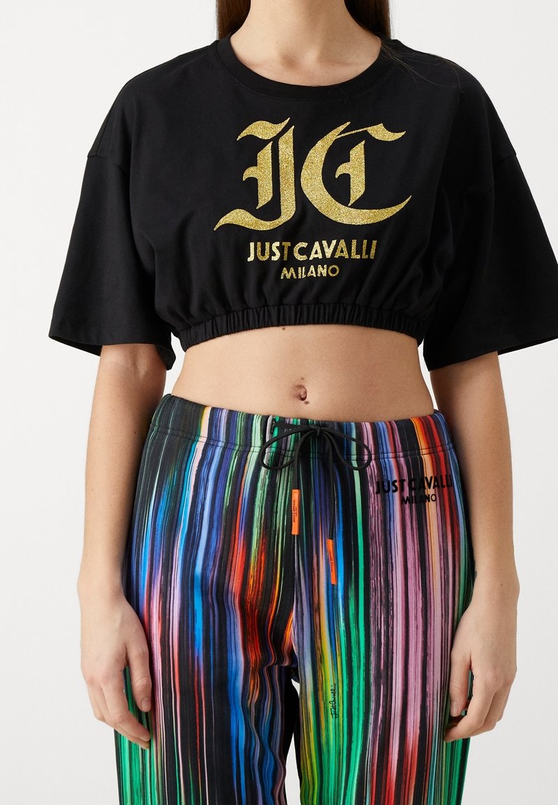 Mustavihreä cropped t-paita, jossa on kultaglitterinen logo "JUST CAVALLI MILANO", yhdistettynä monivärisiin raidallisiin housuihin, joissa on nyörikiinnitysvyötärö.
