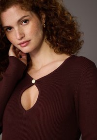 Bordeauxrotes, geripptes Oberteil mit einem Ausschnitt in Tropfenform am Ausschnitt, verziert mit einem goldenen Knopf. Lange Ärmel aus strukturiertem Stoff.