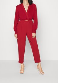 Mono rojo con mangas largas, escote en V profundo, cintura ceñida acentuada por un cinturón dorado y piernas ajustadas. Combinado con sandalias de tacón alto nude.