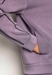 Persona vistiendo un sudadera lavanda con detalle de cremallera negra en la manga, con la mano parcialmente dentro del bolsillo delantero.