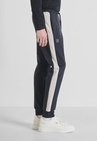 Marine joggerbroek met lichtbeige zijstrepen, elastische tailleband en trekkoord. Bevat een logo patch en geribbelde boorden.