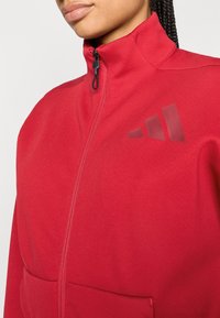 Červená bunda na zips s vysokým golierom, vyrobená z hladkej tkaniny; má čierny zips a nenápadné logo s tromi pruhmi na hrudi.