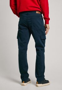 Pantalones cargo azul marino de algodón con bolsillos laterales, corte recto y una etiqueta de cuero en la parte trasera. Llevados con zapatillas oscuras.