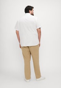 Cămașă albă cu mâneci scurte și tiv curbat, asortată cu pantaloni bej deschis și adidași albi. Viziunea din spate arată o croială lejeră și un design minimalist.