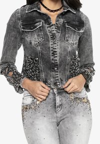 Harmaa farkkutakki, jossa koristeelliset pitsi- ja helmikoristelut hihoissa. Takissa on napitus ja kaksi rintataskua.