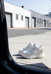 Baskets blanches épaisses avec des fleurs décoratives sur le côté, placées sur un trottoir en ville près des bâtiments industriels en plein jour.