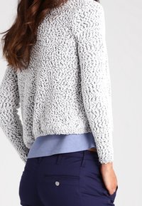 Veste gris clair texturée avec une surface en peluche. Elle a des manches longues et une coupe ample, portée par-dessus une chemise bleue et un pantalon bleu marine.