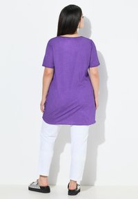 Tunique violette en tissu léger avec des manches courtes, associée à un pantalon blanc et des chaussures avec des accents argentés, vue de dos.