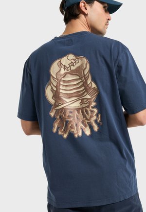 T-shirt en coton bleu marine avec un grand motif doré de pancakes empilés avec du sirop et une fourchette au dos. Manches courtes, coupe décontractée.
