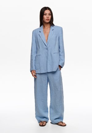 Mujer de pie vistiendo un blazer grande de rayas azul claro y pantalones de pierna ancha con sandalias marrones sobre fondo blanco.