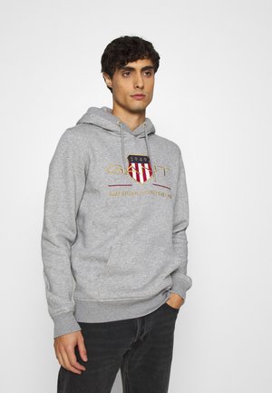 Felpa grigia in materiale morbido, con un logo frontale rosso e oro, cappuccio con lacci e polsini a coste. Indossata con jeans scuri.