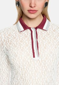 Spitzen-Poloshirt mit einem kastanienbraunen Kragen, der weiße und graue Streifen aufweist. Vorderseite mit Knöpfen und einem strukturierten Stoff mit einem filigranen Muster.