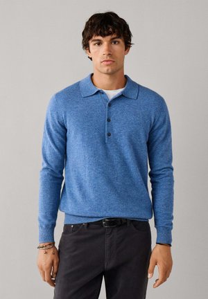 Joven con cabello rizado oscuro, vestido con un jersey polo azul de manga larga sobre una camisa blanca y pantalones negros frente a un fondo neutro.