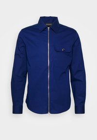 Camisa navy de botones, hecha de una mezcla de algodón, con un cierre de cremallera plateada y un solo bolsillo en el pecho con cierre a presión. Cuello estructurado y mangas largas.
