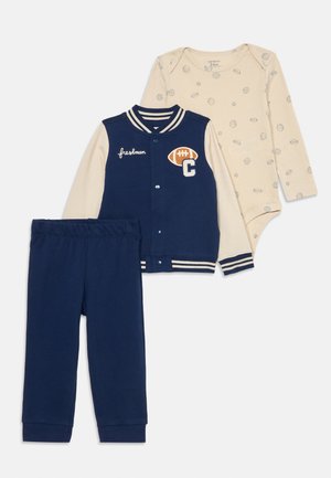 Casaco varsity azul-marinho com punhos listrados em branco, macacão bege com padrões de bolas e calças azuis-marinho combinando. Material de algodão suave.