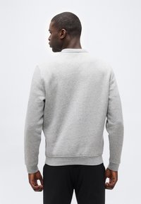 Άνδρας με την πλάτη προς τα εμπρός, φοράει ανοιχτογκρι μακρυμάνικη μπλούζα και μαύρο παντελόνι, με φόντο ένα απλό λευκό σκηνικό.