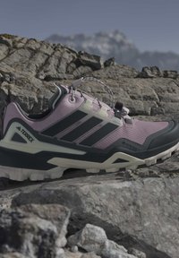 Лилави и черни туристически обувки Adidas Terrex Gore-Tex, почиващи върху груби сиви скали с далечни размазани планини под ясно небе.