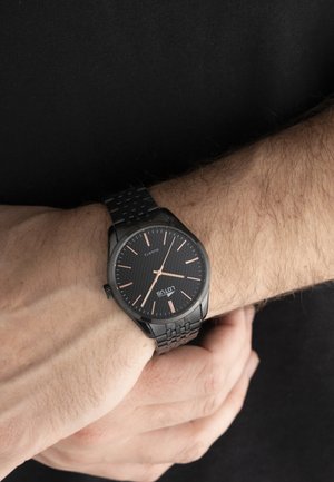 Crni metalni ručni sat s ružičasto zlatnim kazaljkama i oznakama, nošen na lijevoj ruci na crnoj pozadini.