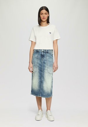 Weißes Baumwoll-T-Shirt mit einem kleinen blauen Herzlogo, kombiniert mit einem hellblauen Jeans-Midikleid mit vorderen Taschen und einem Verblassungs-Effekt.