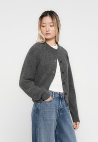 Junge Frau mit halb-aufgebleichtem Haar, die eine graue Strickjacke mit Knöpfen über einem weißen Top und lockeren blauen Jeans trägt, posiert mit einer Hand in der Tasche.
