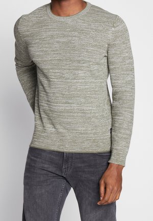 Muž nosící světle šedý svetr s strukturovaným vzorem a dlouhým rukávem a tmavě šedé džíny, stojící před neutrálním světlým pozadím.