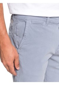 Detailní záběr na ruku osoby odpočívající vedle světle modrých volnočasových kalhot s poutky na opasek a předními kapsami, oblečenou v bílém tričku.