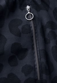 Tissu noir avec un motif animalier subtil, doté d'une fermeture éclair argentée avec un tirette circulaire. Texture douce, vue en gros plan.