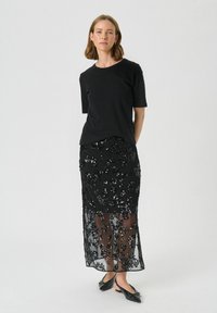 Haut noir à manches courtes associé à une longue jupe noire en tissu transparent ornée de motifs floraux en sequins complexes. Des ballerines noires complètent le look.