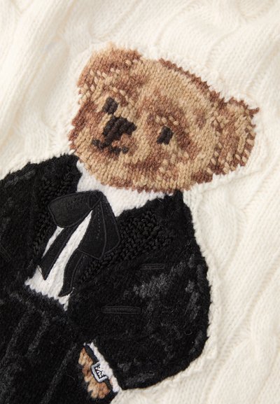 Maglione lavorato a maglia con un orso marrone in smoking su uno sfondo crema. Motivo a trecce testurizzate con dettagli in smoking neri.