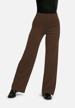Person iført højtaljede, vidde-ben-brune bukser med sorte loafers og en sort top, stående med benene let adskilt på en hvid baggrund.