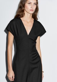 Svart klänning med V-ringning och korta ärmar, med en figursydd silhuett och slät textur. Minimalistisk design utan synliga mönster eller dekorationer.