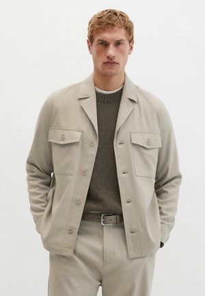 Blondyn w beżowej, zapinanej na guziki kurtce na szarym dzianinowym swetrze, beżowych spodniach i pasującym pasku, stojący z rękami w kieszeniach.