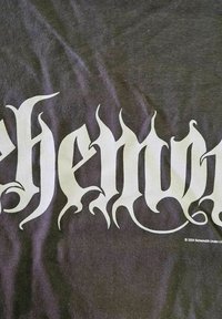 Mørkegrå t-skjorte med stor, hvit stilisert tekst som leser "behemoth" med flammeaktige detaljer. Myk tekstur synlig.