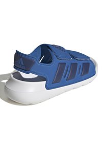 Modra atletska sandal s teksturiranim zgornjim delom, nastavljivim paščkom in belo oblazinjeno podplatom. Ikoničen detajl treh črt na strani.