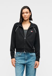 Μαύρο μπουφάν με φερμουάρ και λευκές λεπτομέρειες, κατασκευασμένο από λείο ύφασμα. Διαθέτει μικρό λογότυπο στο αριστερό στήθος και συνδυάζεται με ανοιχτόχρωμα μπλε τζιν.