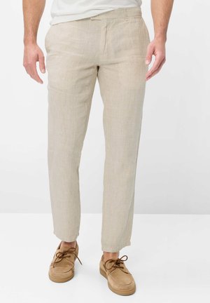 Man draagt beige linnen broek en lichtbruine suède veterschoenen, staand tegen een witte achtergrond.