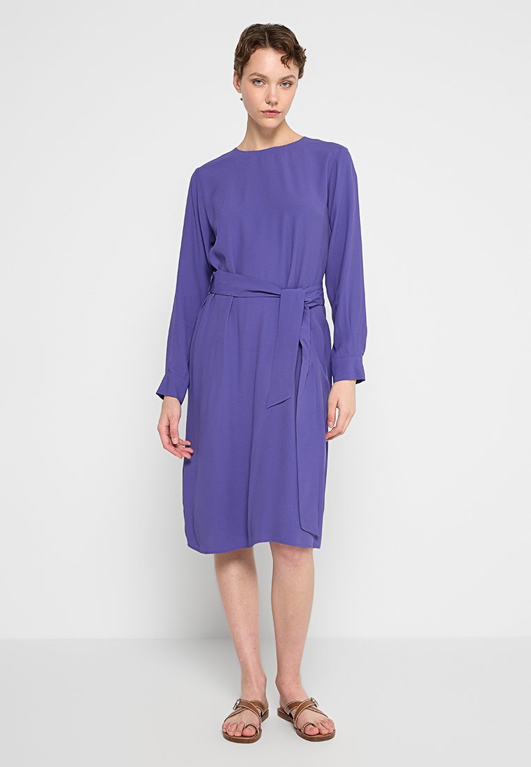 Langes lila Kleid mit langen Ärmeln aus glattem Stoff, mit einem Gürteldetail in der Taille und seitlichen Taschen, knielanger Schnitt.
